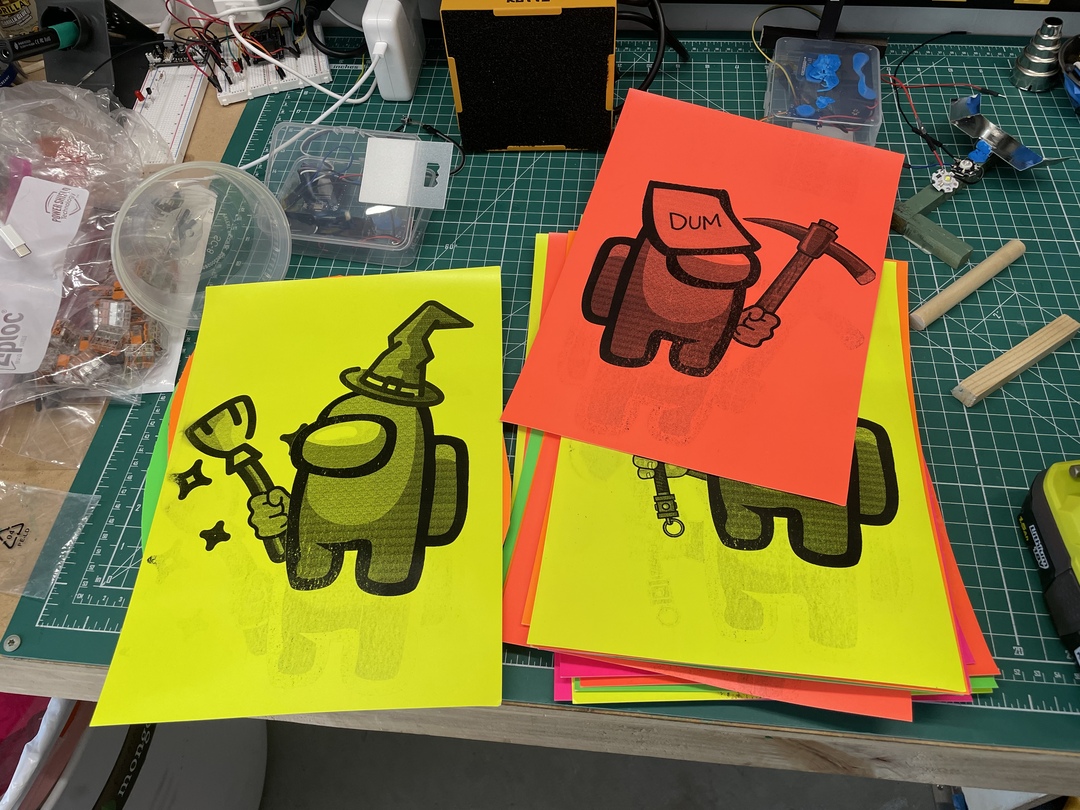 Brightly colored Among Us characters with tools, printed on fluorescent paper, scattered on a workbench.