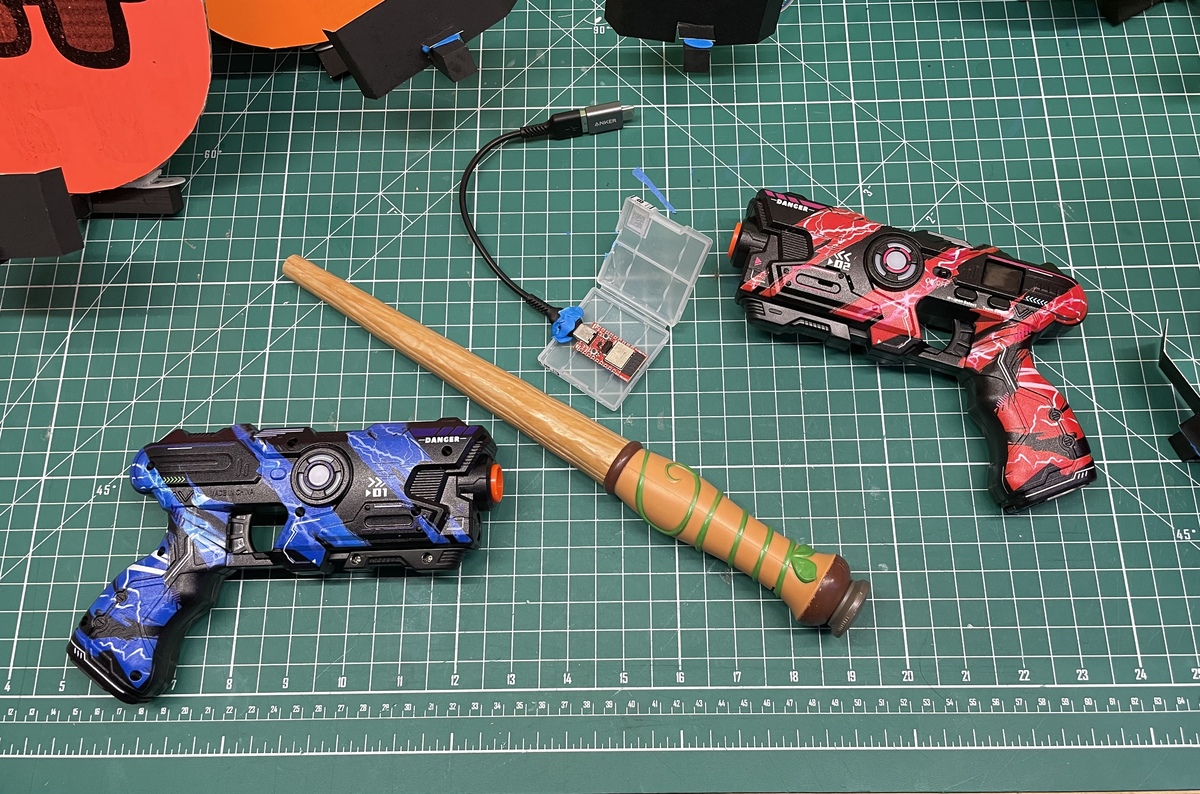 Two toy laser tag pistols in blue and red lightning designs and a wooden wand with tan grip wrappings, arranged on a green cutting mat. A small clear plastic case containing electronic components and a USB cable is also visible.